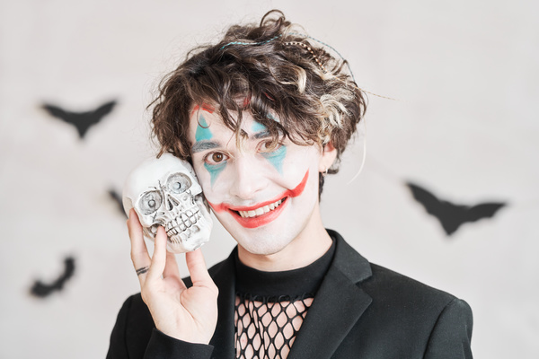 Man with Joker Makeup Stands against Background Bats and Holds Skull to His Face